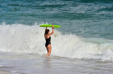 Slim girl with a surfboard in a swimsuit on the beach in front of a big wave