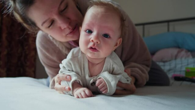 A tender moment between a baby and mum, who is gently kissing the babys forehead. The baby, with striking blue eyes and wisps of light hair, a thread of saliva visible on their chin.