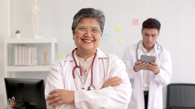Asian professional senior female doctor smiles confidently looking camera, experienced medical professional white coat collaborates work with young specialist male colleague focus on tablet in clinic