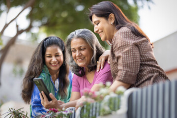 Three female friends laughing at something on a mobile phone while standing together outside in a back yard