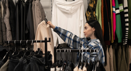 Brunette woman in fashion store choosing clothing items to buy. Girl near Racks of stylish clothes with sweater in hands, examining before making a decision.