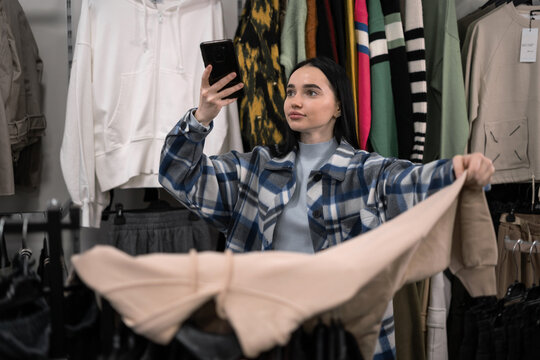 A woman shopping in a shopping center chooses clothes in a fashion store and takes a photo on her smartphone.