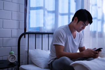 Fototapeta premium Asian teenager boy scroll mobile phone, search and online watching online movie after chat message and playing internet with his friend in bedroom of a dimly light