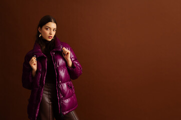 Fashionable confident woman wearing trendy purple puffer down  jacket, posing on brown background....