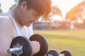 Young asian plump boy doing exercise with lifting heavy dumbbells in outdoor park in the sunset...