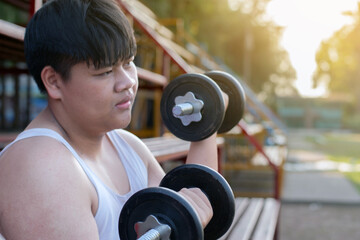 Young asian plump boy doing exercise with lifting heavy dumbbells in outdoor park in the sunset...