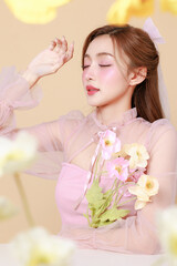 Young cute Asian woman in a pink elegant dress, Korean style makeup, moisturized, smooth, perfect skin with flowers on a beige background. Facial treatment, Cosmetology, plastic surgery.