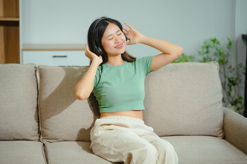 young Asian woman relaxes on the sofa, listening to music. She enjoys a moment of tranquility,...