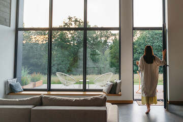 Woman standing with cup by the fireplace near the window at modern house on nature during summer...