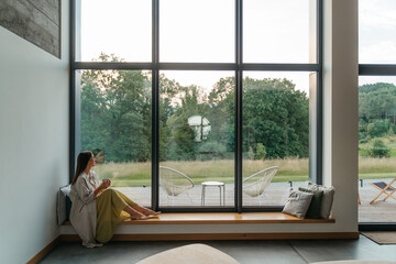 Young woman resting at beautiful country house or hotel, sitting on the window sill enjoying beautiful view on pine forest. Concept of solitude and recreation on nature