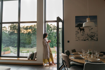 Woman standing with cup by the fireplace near the window at modern house on nature during summer...