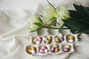 Happy Easter! Stylish easter eggs in tray and tulips on rustic table flat lay. Natural candy chocolate eggs with spring flowers. Easter hunt, space for text