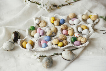 Stylish colorful easter eggs in tray and cherry blossoms on rustic table with linen cloth. Happy Easter! Natural candy chocolate eggs with spring flowers. Easter hunt