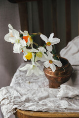Beautiful daffodils in vintage vase on rustic wooden chair with linen cloth. Countryside floral still life. Rural moody spring flowers composition. Easter banner
