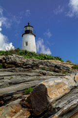 Fototapeta na wymiar Pemaquid Point Lighthouse in Bristol Maine sits atop rugged rock layers on a bright summer day dotted with puffy clouds
