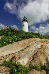Pemaquid Point Lighthouse in Bristol Maine sits atop rugged rock layers on a bright summer day dotted with puffy clouds