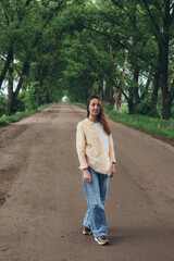 A stylish girl, a woman stands in a tunnel of green trees. Warm summer day. The wind blows the girl's hair and clothes. Girl looking at the camera