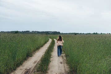 A girl, a woman walks into the distance along the road among a field of green wheat. Summer cloudy day. Hair blows in the wind