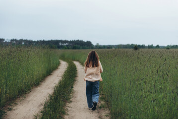A girl, a woman walks into the distance along the road among a field of green wheat. Summer cloudy day. Hair blows in the wind