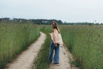 A girl, a woman walks into the distance along the road among a field of green wheat. Summer cloudy...