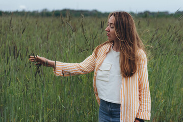 A girl, a woman stands in the middle of a field of green wheat. Touches ears of wheat with his hands. Summer cloudy day.
