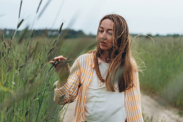 A girl, a woman stands in the middle of a field of green wheat. Touches ears of wheat with his hands. Summer cloudy day. The wind blows the girl's hair.