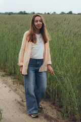 A girl, a woman stands on the road in the middle of a field of green wheat.