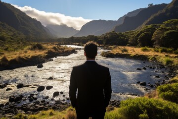 A man in a suit stands by a river, gazing at snowy mountains in the distance under a clear blue sky. He looks contemplative and relaxed, hands in pockets.