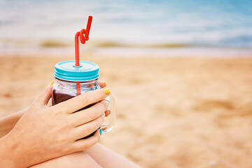 Glass jar with a drink and a straw in women's hands. Female sitting on the sea beach with an alcoholic cocktail. Vacation on tropical coast. Photo card with copy space.