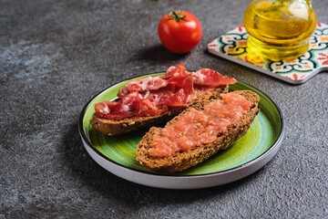 Spanish breakfast, two open tostada sandwiches with grated tomato and salted thinly sliced pork on dark bread on a green ceramic plate on a dark concrete background.