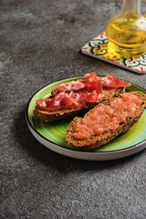 Spanish breakfast, two open tostada sandwiches with grated tomato and salted thinly sliced pork on dark bread on a green ceramic plate on a dark concrete background.