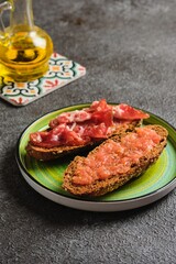 Spanish breakfast, two open tostada sandwiches with grated tomato and salted thinly sliced pork on dark bread on a green ceramic plate on a dark concrete background.