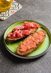 Spanish breakfast, two open tostada sandwiches with grated tomato and salted thinly sliced pork on dark bread on a green ceramic plate on a dark concrete background.