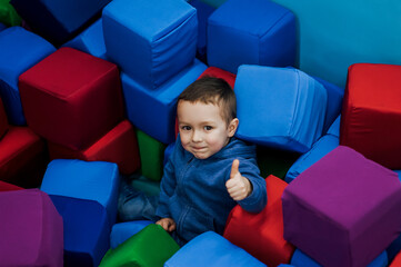Little boy, happy smiling child lying down playing with soft colored cubes in playroom, showing...