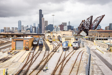 Diesel locomotives at a suburban train depot on a cloudy day. Downtown Chicago Skyline and an open draw bridge are in background.