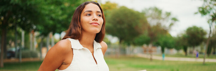 Lovely tanned woman with long brown hair wearing white top sitting relaxed on park bench looking away, Panorama. Life style concept