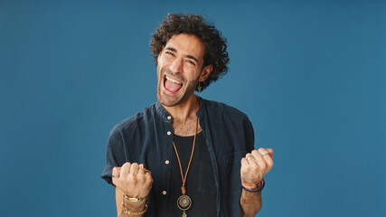Happy attractive man with curly hair, wearing blue shirt, looking at camera surprised wow raises his hands celebrating victory isolated on blue background in studio - obrazy, fototapety, plakaty