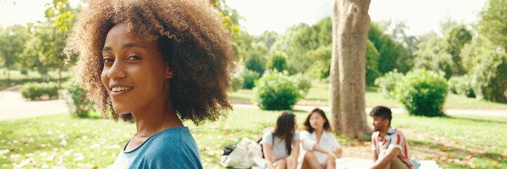 Portrait of young smiling woman with curly hair wearing blue t-shirt posing for the camera in the park, Panorama. Picnic on summer day outdoors her friends sitting in distance blurred on background - obrazy, fototapety, plakaty