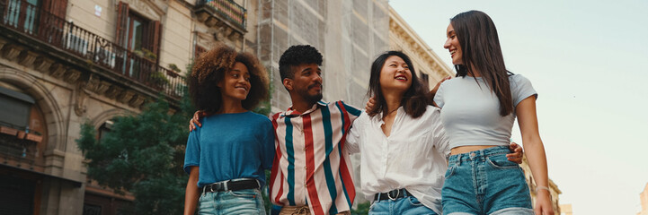 Happy multiethnic young people walk embracing on summer day outdoors, Panorama. Group of friends are talking and laughing merrily while walking along on the street