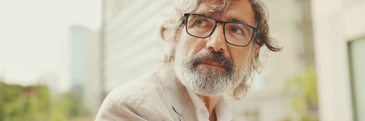 Clouse up portrait of mature businessman with beard in eyeglasses wearing gray pondering jacket...