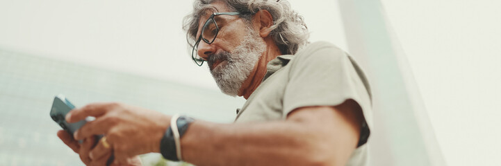 Friendly middle aged man with gray hair and beard looking at map trying to find his way using his mobile phone, Panorama. Mature gentleman in eyeglasses using map app in cellphone outdoors