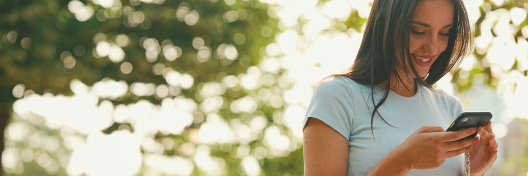 Young Beautiful Smiling Woman With Long Brown Hair Wearing White Crop Top Sitting In Park Having Picnic On Summer Day Outdoors, Talking With Friends, Using Cellphone, Panorama