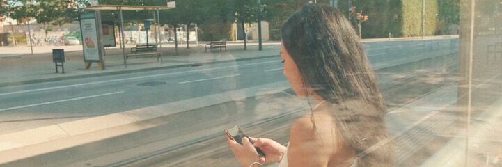 Young woman sits at public transport stop, uses mobile phone, blurry view through the glass, Panorama