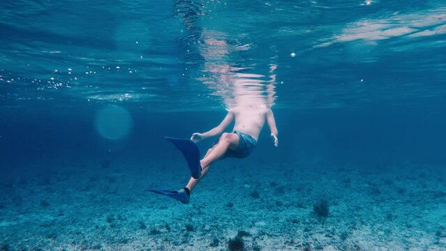 A tall Caucasian guy in a snorkeling mask swims and turns over underwater and swims looking at camera. Guy swims in ocean backwards pushing off with big blue flippers. Pale man on vacation on islands