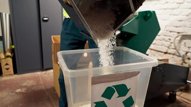 close up A waste recycling plant employee in uniform and headphones pours shredded plastic caps into container for recycling