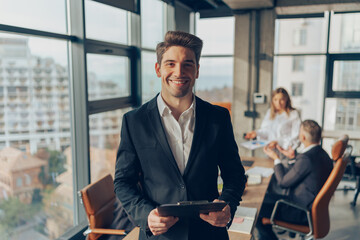 Smiling businessman in suit standing with clipboard at office on colleagues background