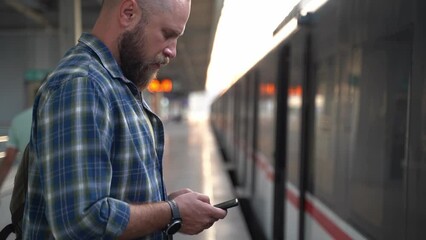 Man with backpack awaiting metro train on railway platform. Adult going to work using gadget smartphone in Public transport. Urban life citizen in morning using subway. Concept of habit in transport