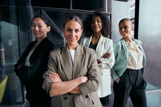 Team portrait of successful happy multiracial business women posing confident inside offices. Group of four diverse attractive female professional in suits with arms crossed looking smiling at camera