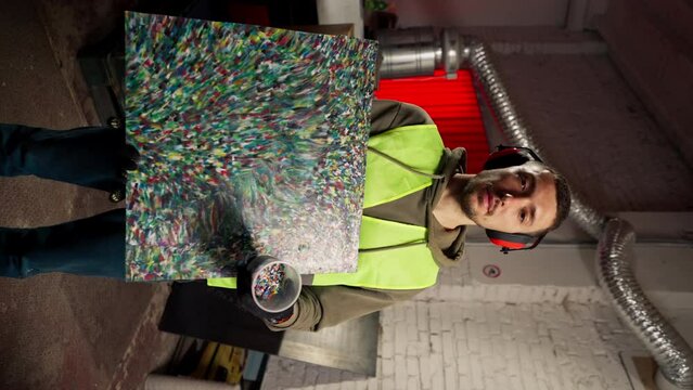vertical video a worker at a waste recycling plant holding a painting made from shredded recycled plastic lids