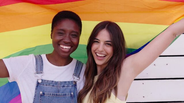 Multiracial beautiful LGBT lesbian loving couple looking smiling at camera hugging affectionate holding rainbow flag high behind them outdoor. Two cheerful young females celebrating pride day together